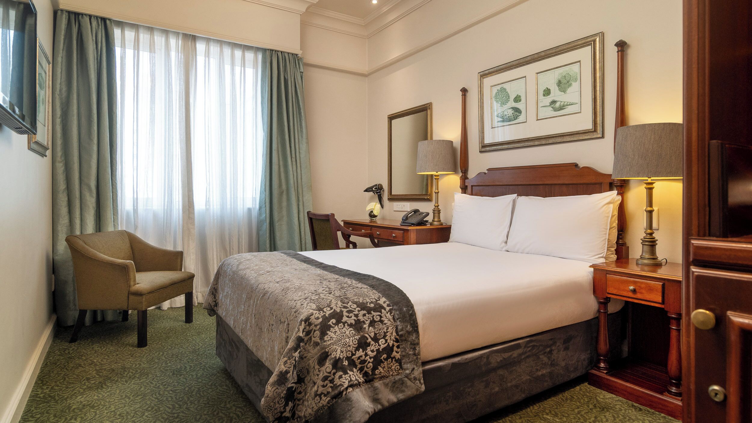A hotel room with a bed, a chair, a desk with a lamp, a telephone, and a framed botanical illustration on the wall.