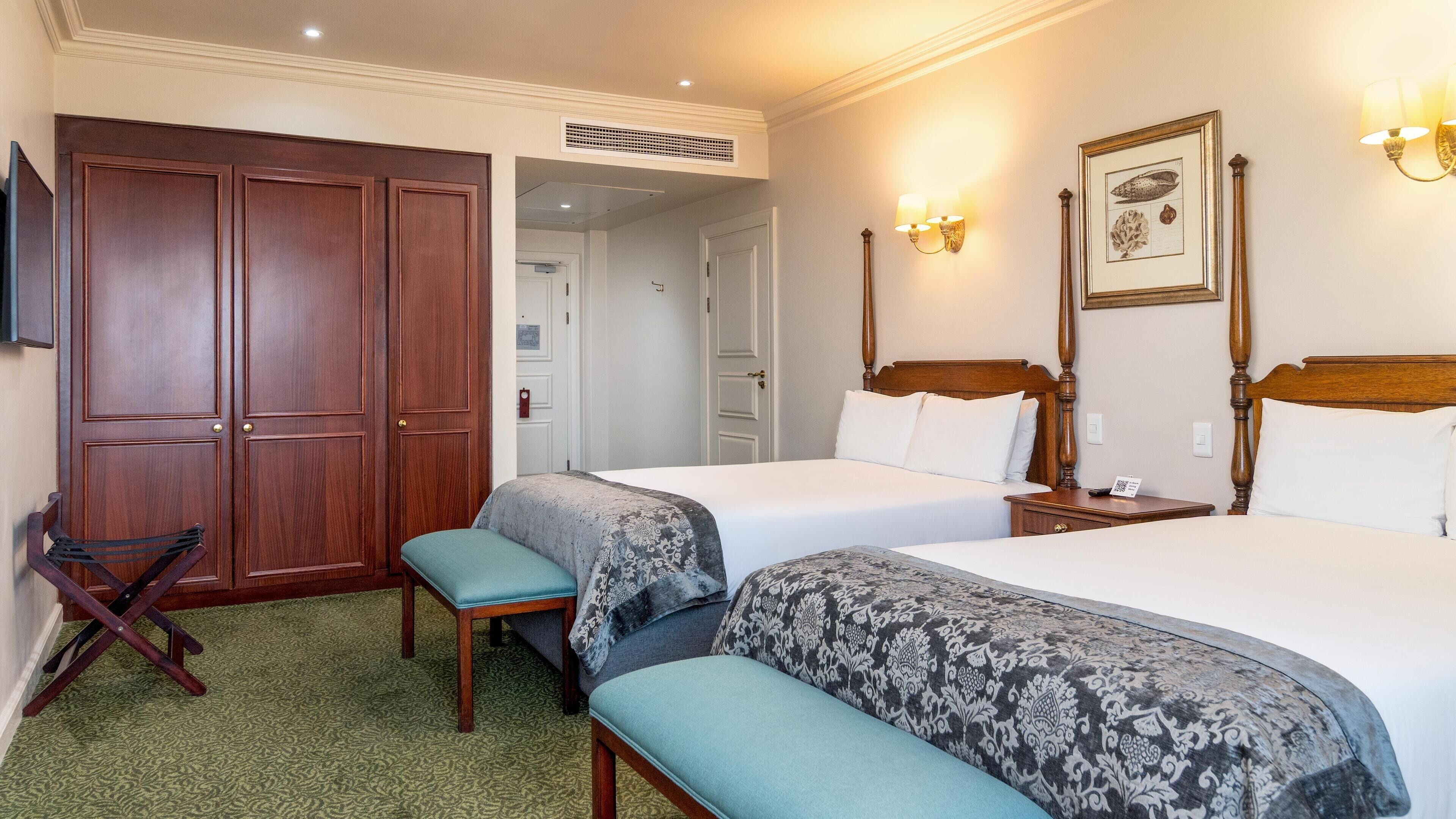A hotel room with two beds, a wooden wardrobe, a bench, and a wall-mounted lamp.