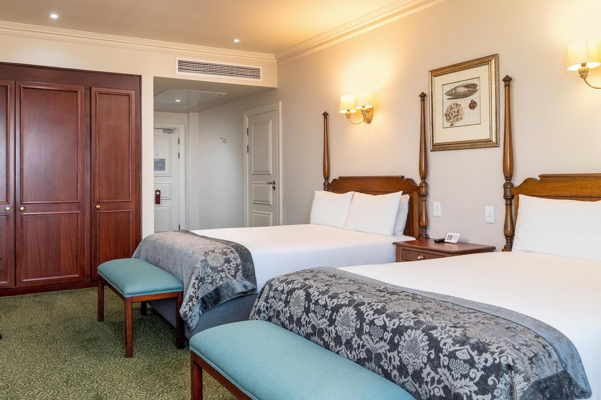 Habitación de hotel con dos camas, un armario de madera, un banco y una lámpara de pared.