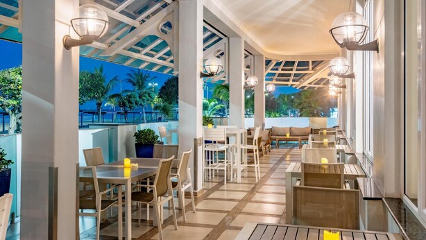 A spacious outdoor dining area with tables and chairs, large windows, and a view of palm trees. - The Edward Hotel (Durban)
