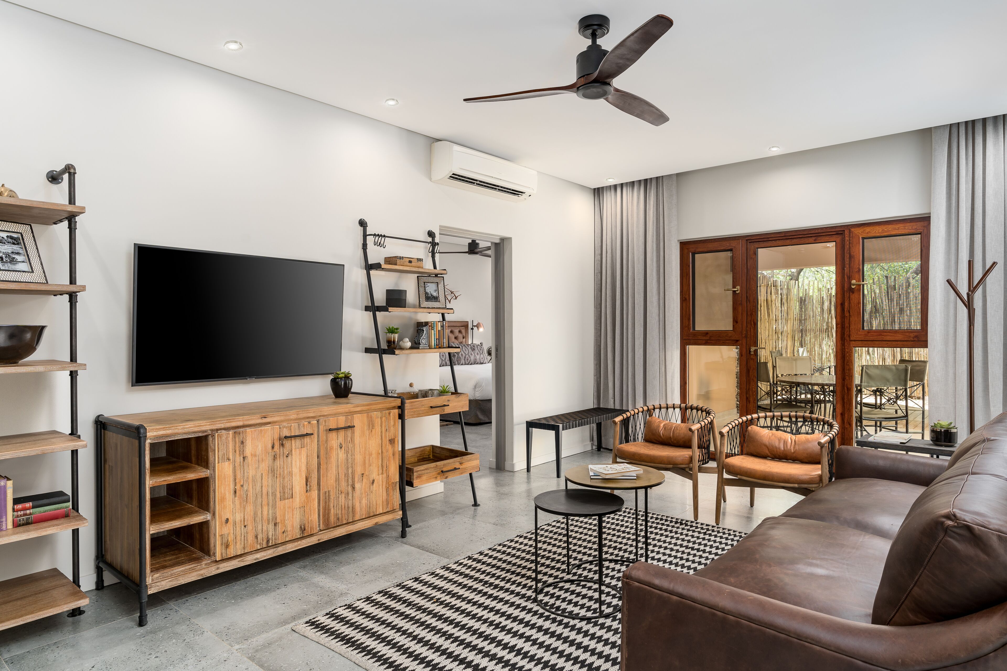 A modern living room with a wooden entertainment unit, a flat-screen TV, a ceiling fan, and a large window with curtains.