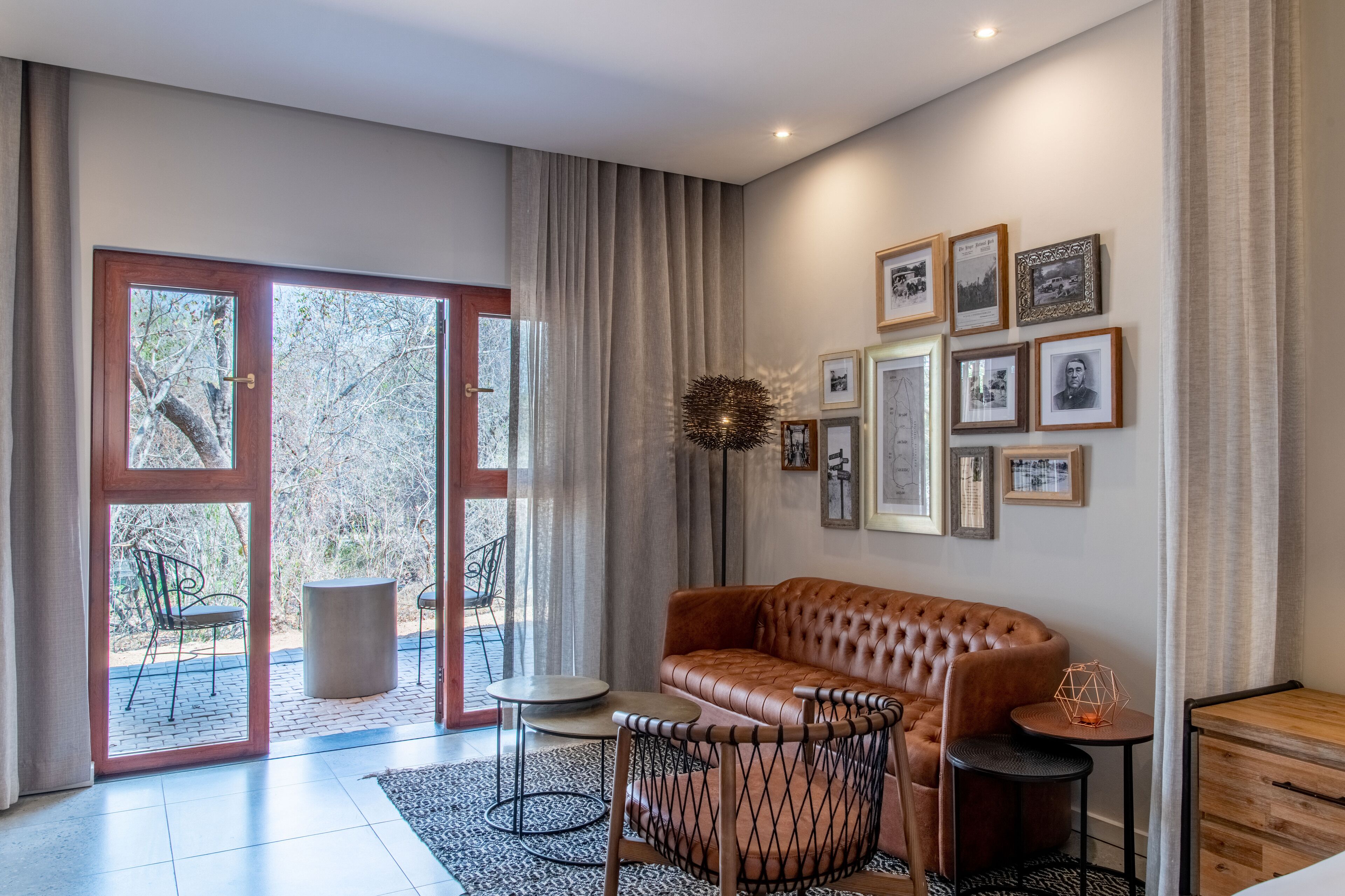 A modern living room with a leather sofa, a dining table, and chairs. There are framed pictures on the wall and a large window with curtains.
