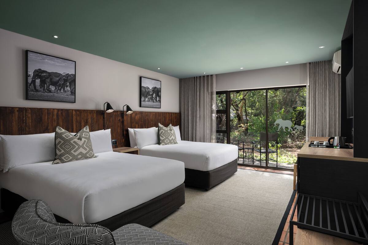 A hotel room with two beds, a desk, and a view of greenery outside.