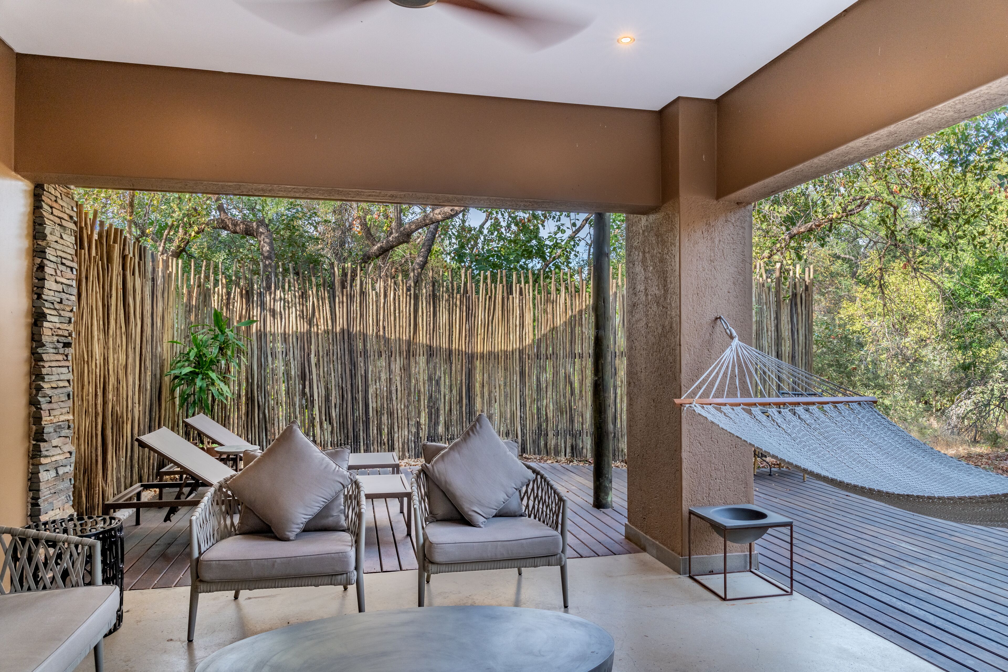 A modern outdoor lounge area with wicker furniture, a hammock, and a view of a bamboo fence and trees.