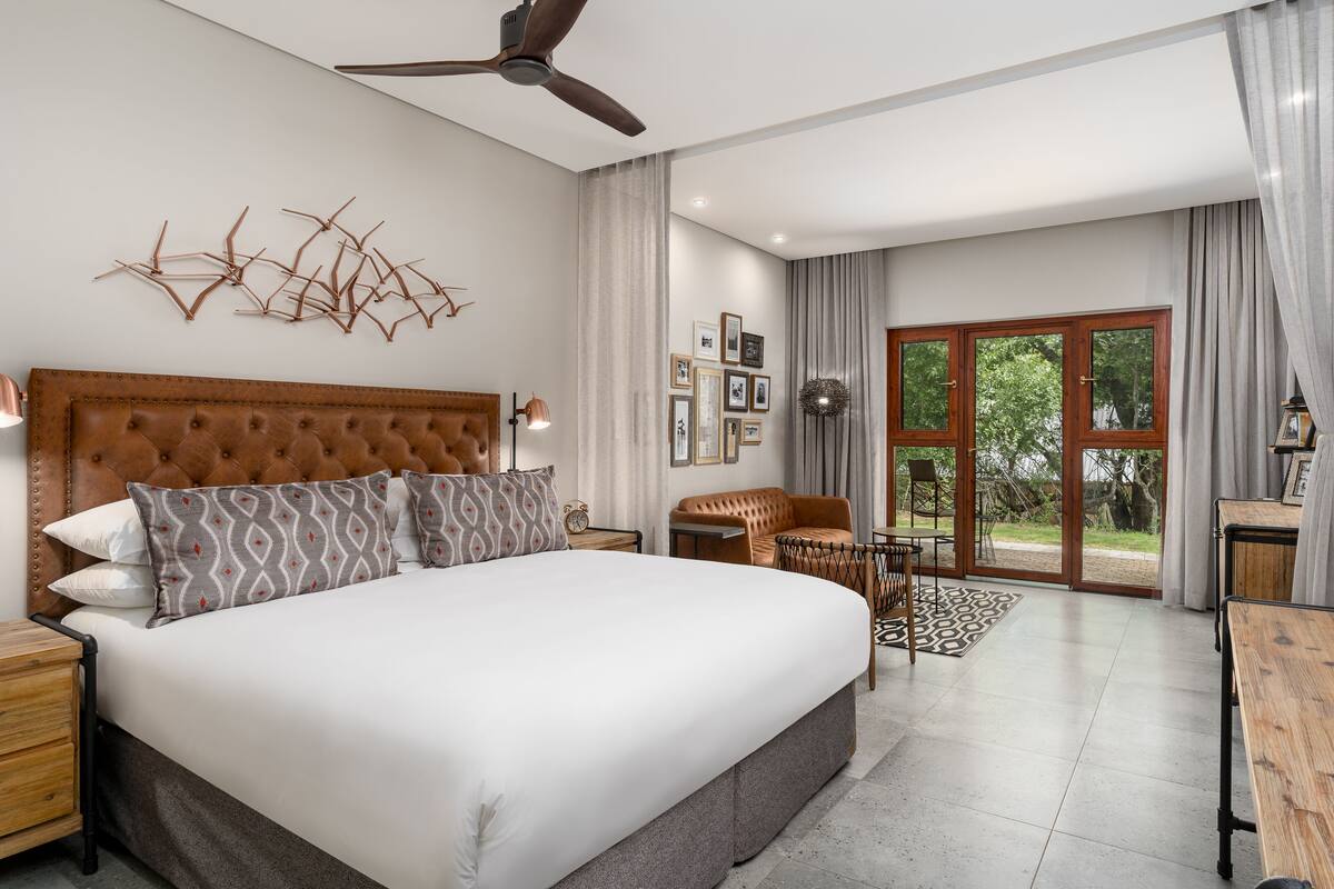 A spacious bedroom with a large bed, a ceiling fan, a seating area with a sofa and chairs, and a view through a glass door to the outdoors.
