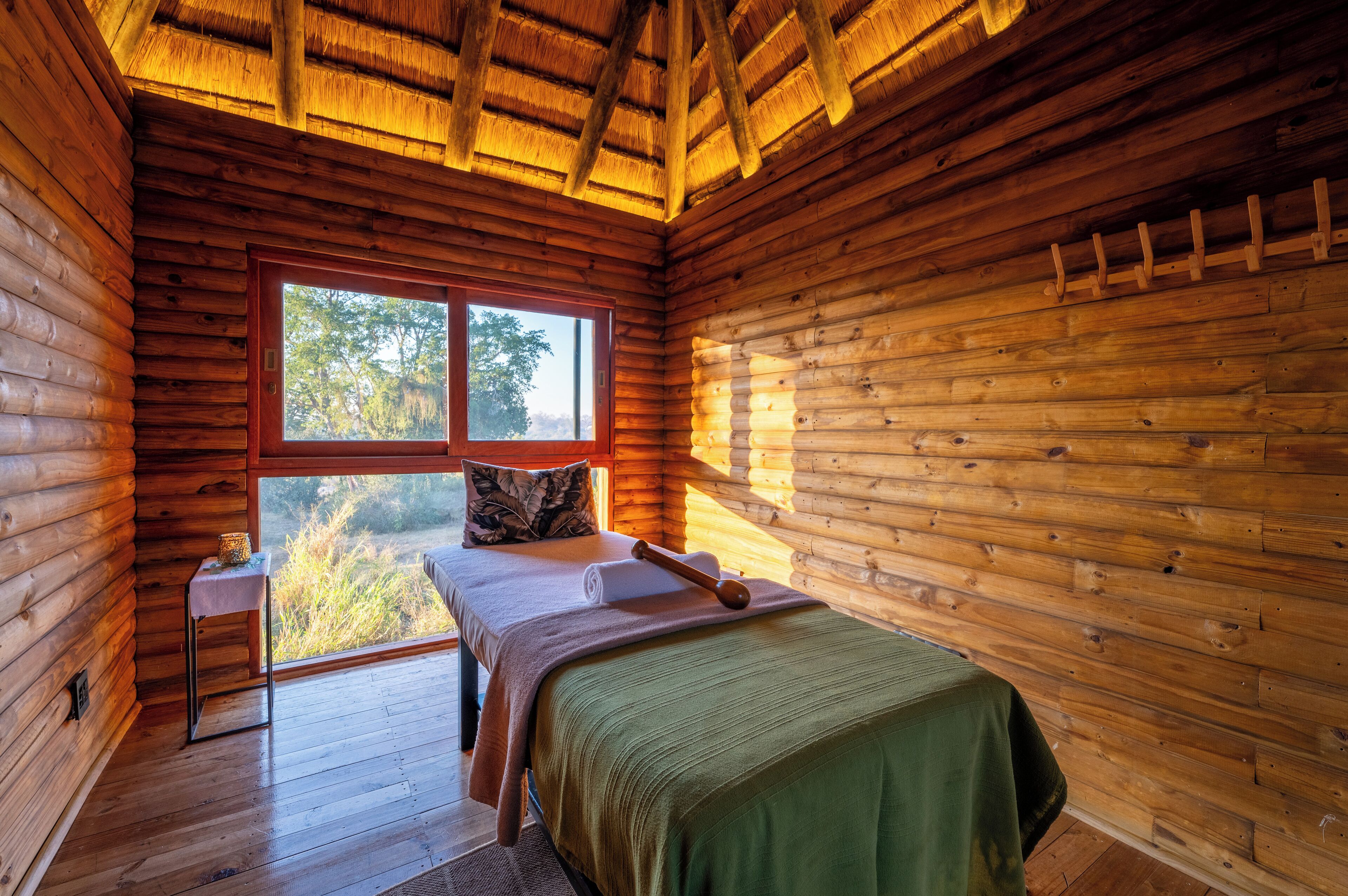 Un dormitorio de cabaña de madera con una cama, una mesita y un ventanal con vistas a árboles.