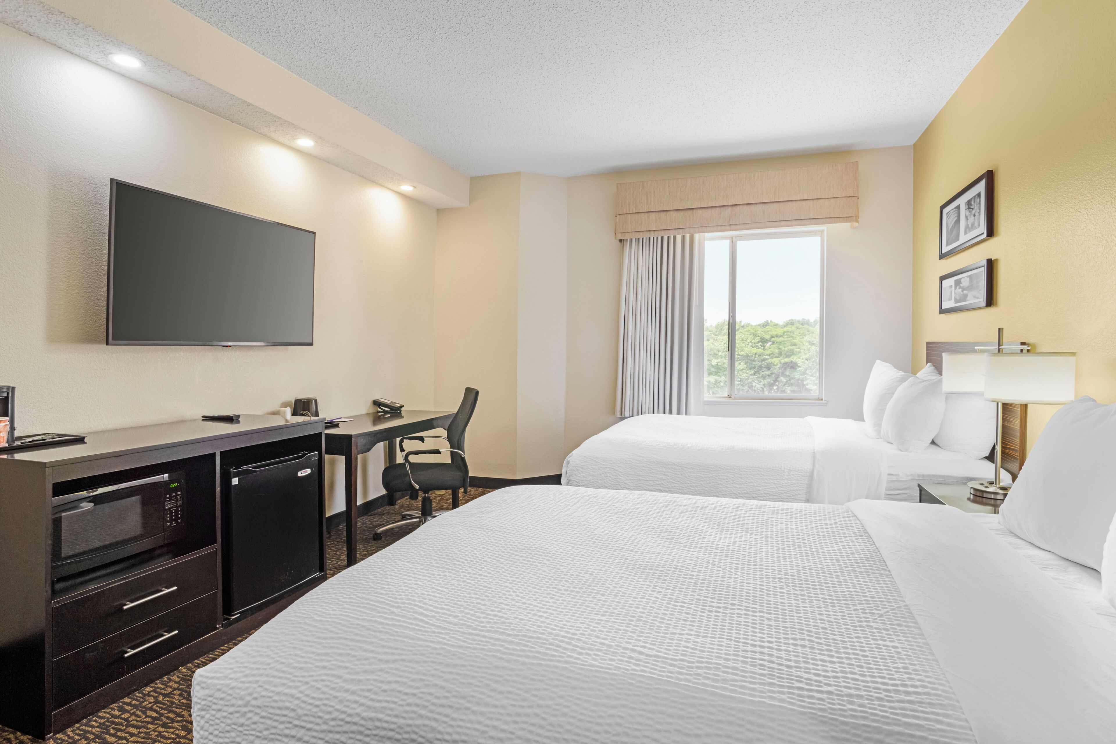 A hotel room with two beds, a desk, a flat-screen TV, a mini-fridge, and a window with curtains.