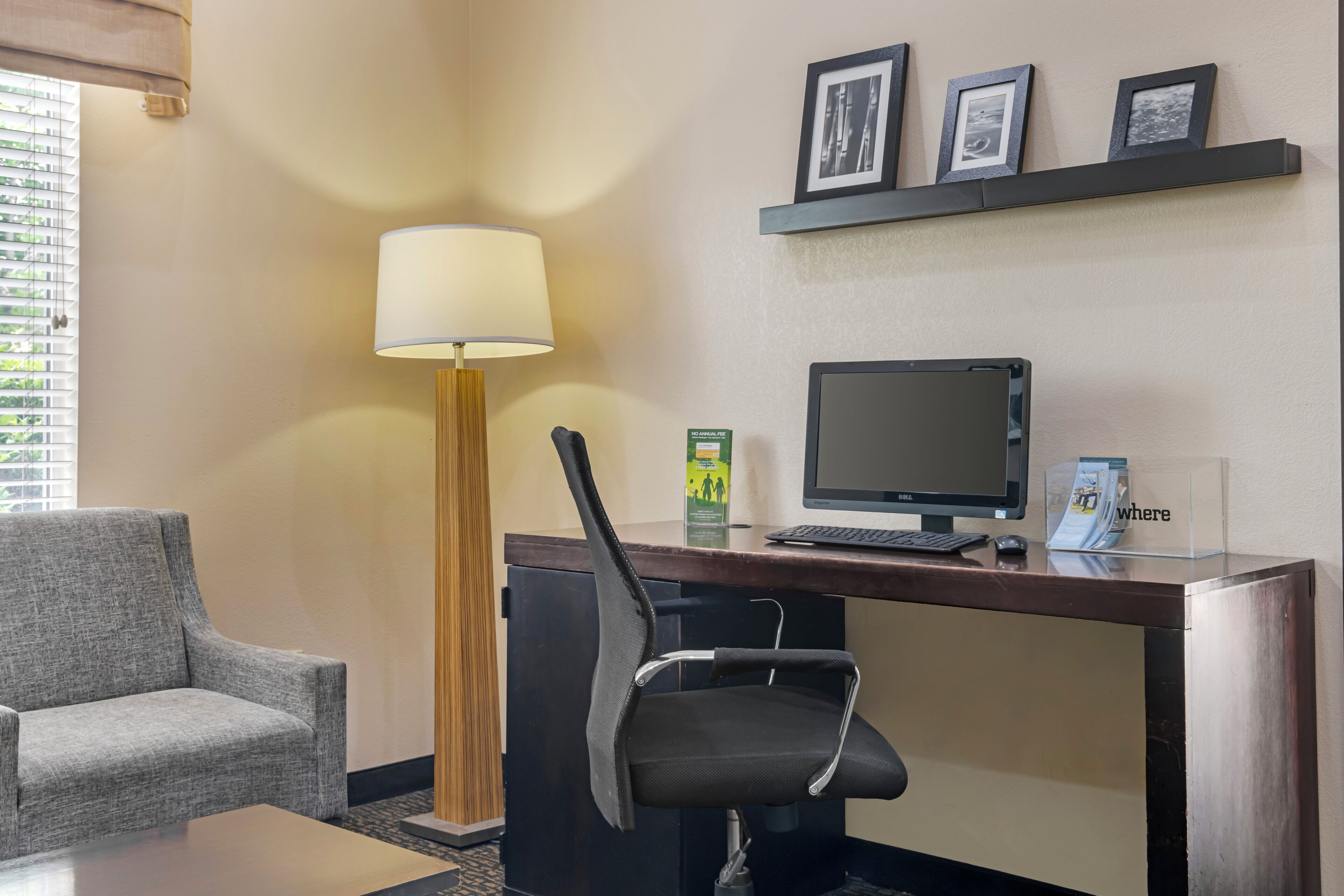 A hotel room with a desk, computer, chair, lamp, and a sofa.