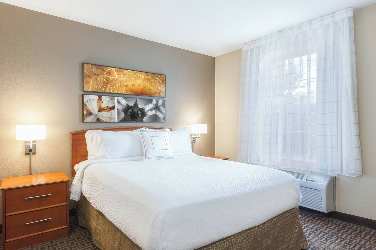 A hotel room with a large bed, two bedside tables, a wall-mounted artwork, and a window with sheer curtains.