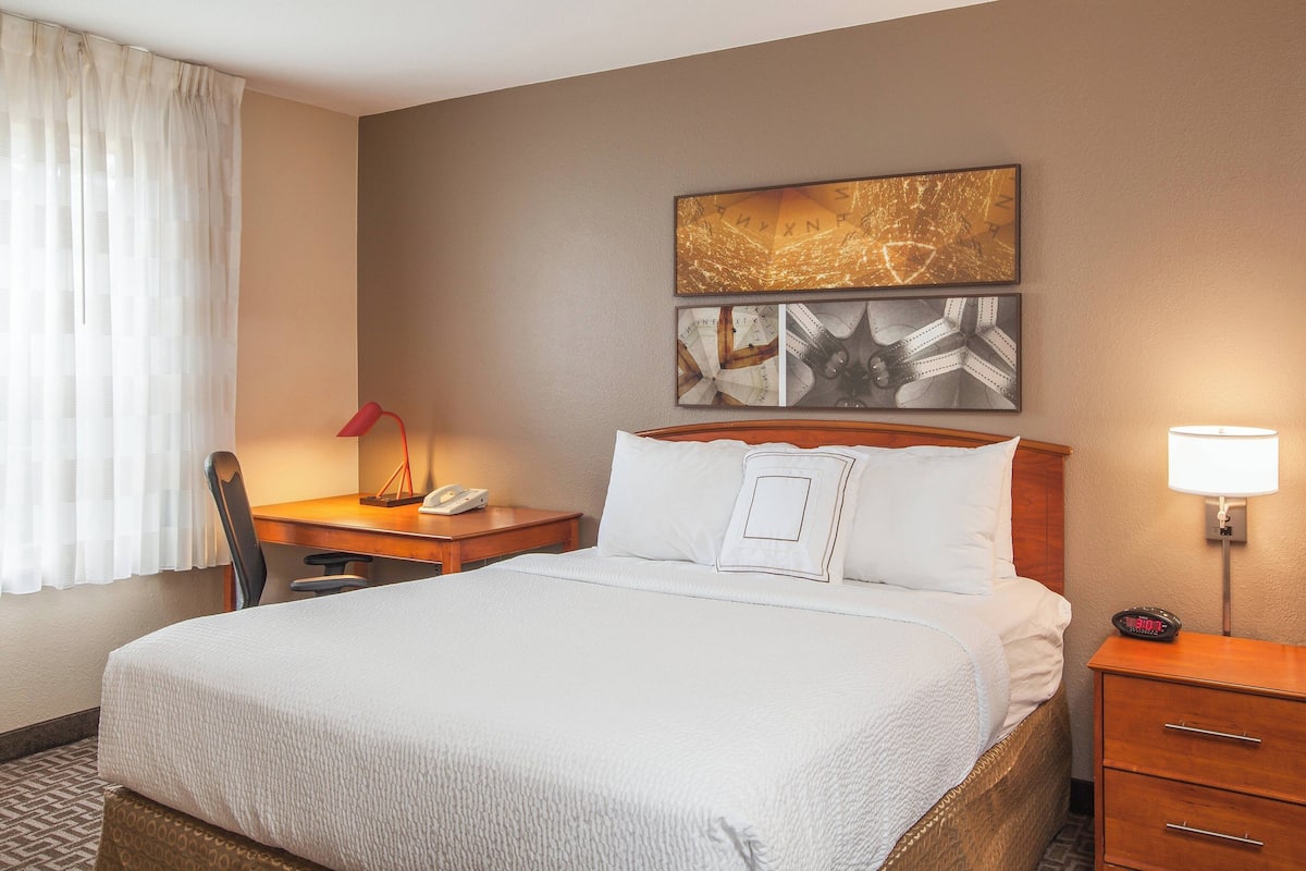 A hotel room with a large bed, a desk with a phone, a chair, a nightstand with a lamp, and artwork on the wall.