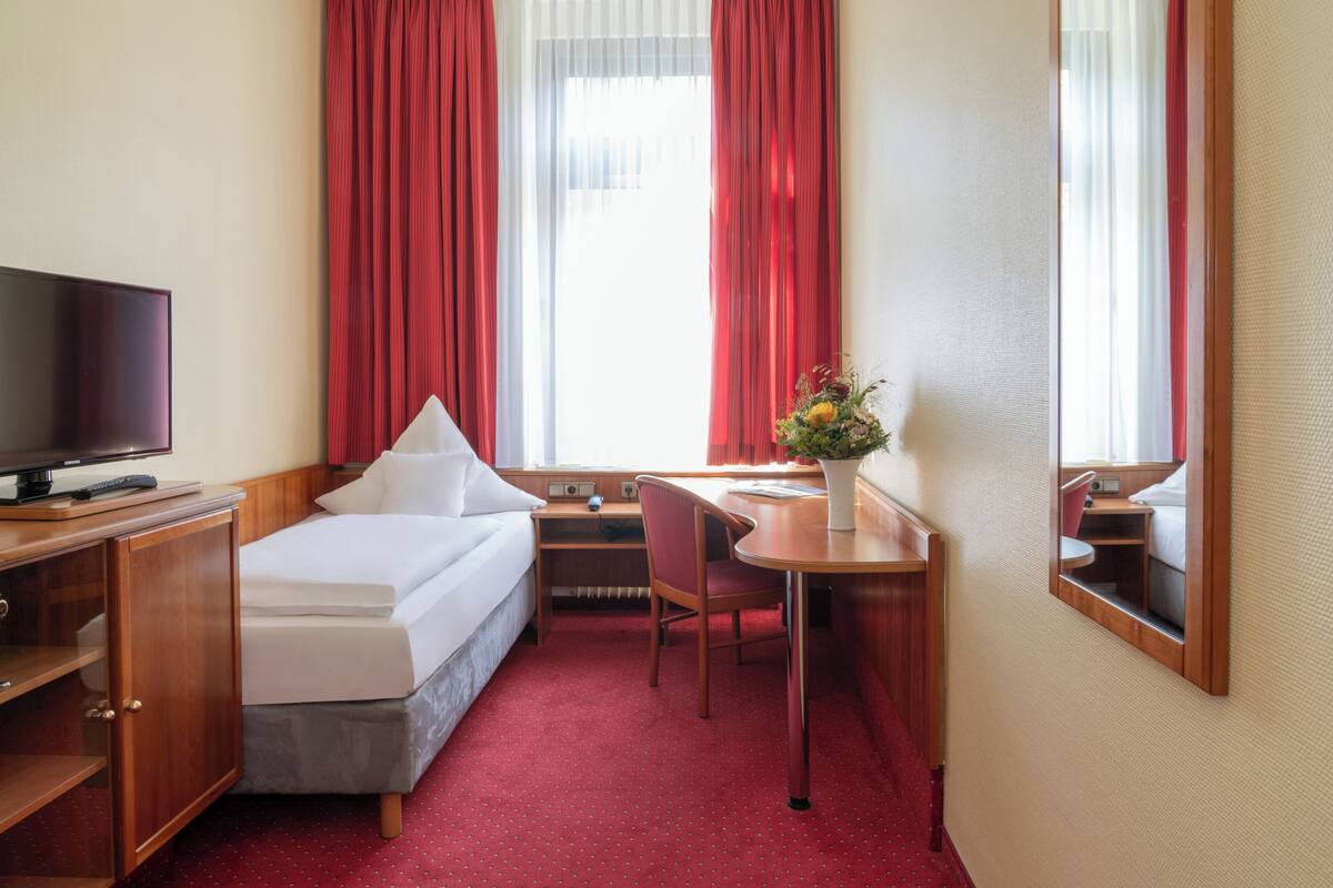 A hotel room with a bed, desk, chair, TV, and a window with curtains.