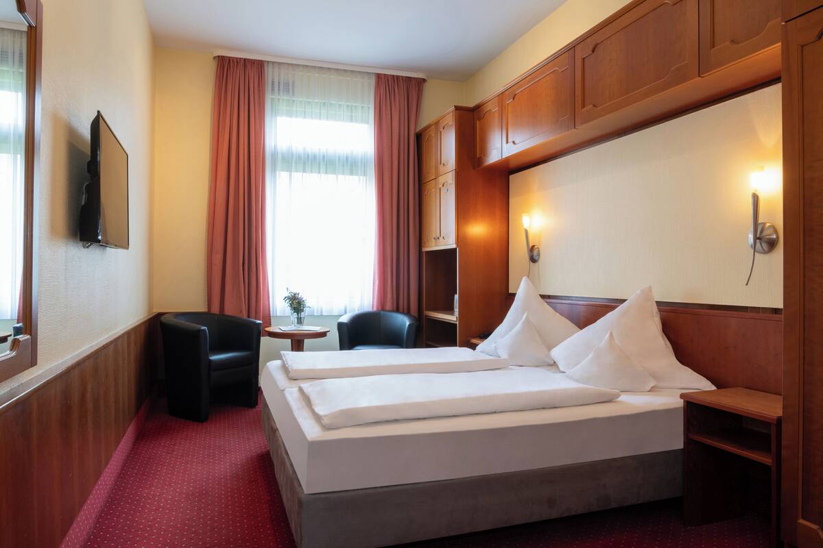 A hotel room with a bed, two bedside tables, a chair, a small table with a vase, and a television mounted on the wall.
