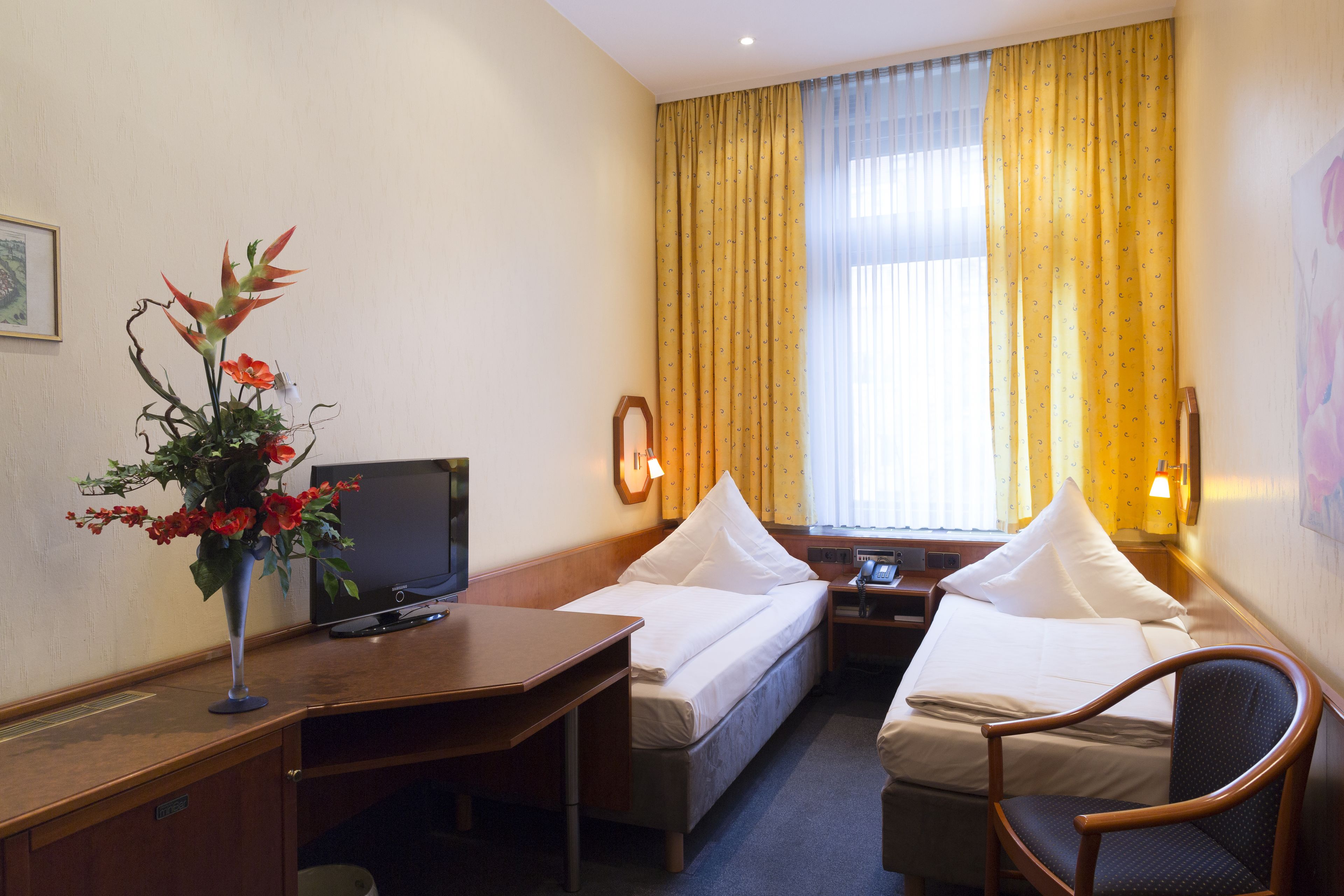 A hotel room with a bed, a desk, a chair, a TV, and a vase with flowers.