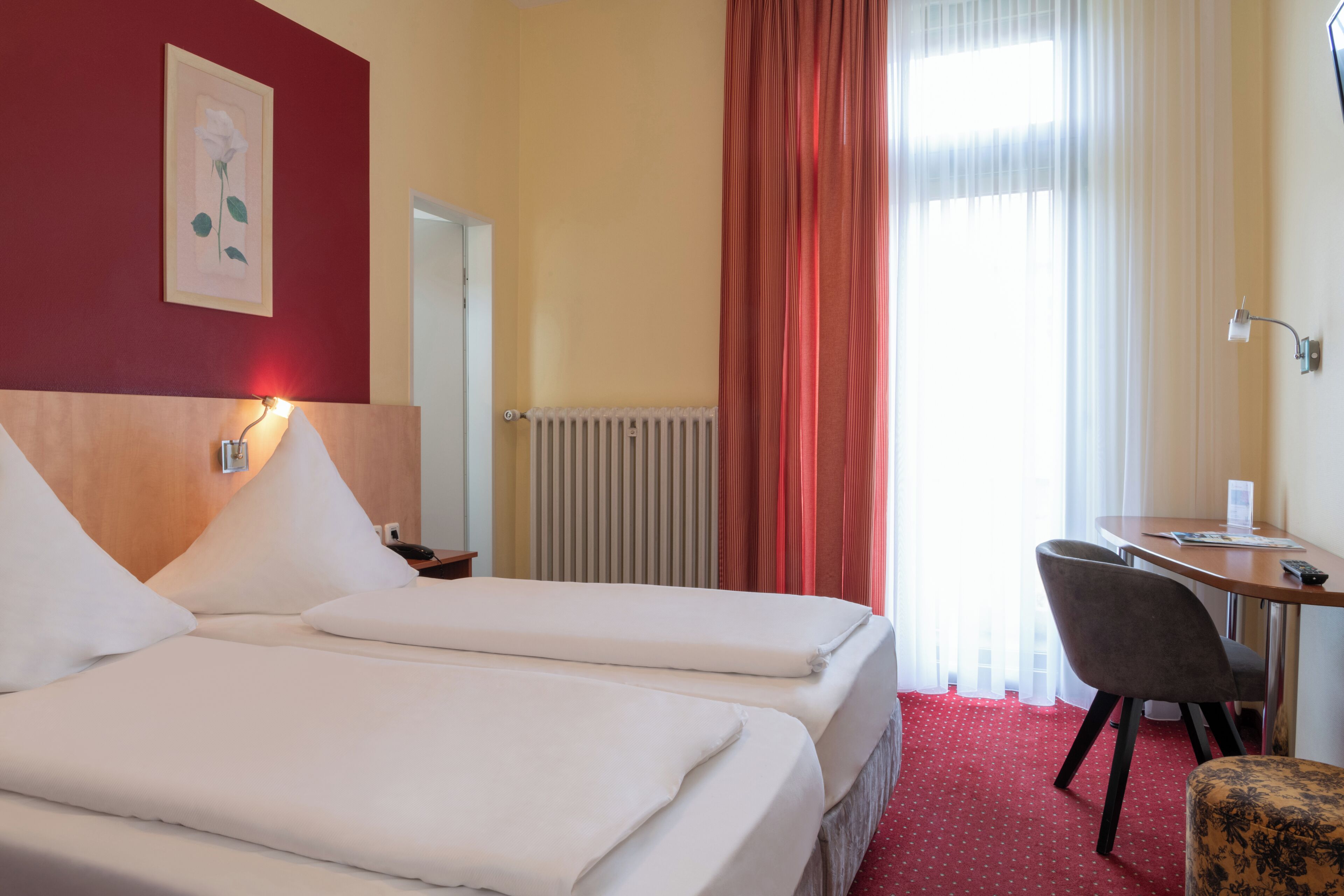 Une chambre d’hôtel avec deux lits, un bureau, une chaise, un radiateur et une fenêtre avec des rideaux.