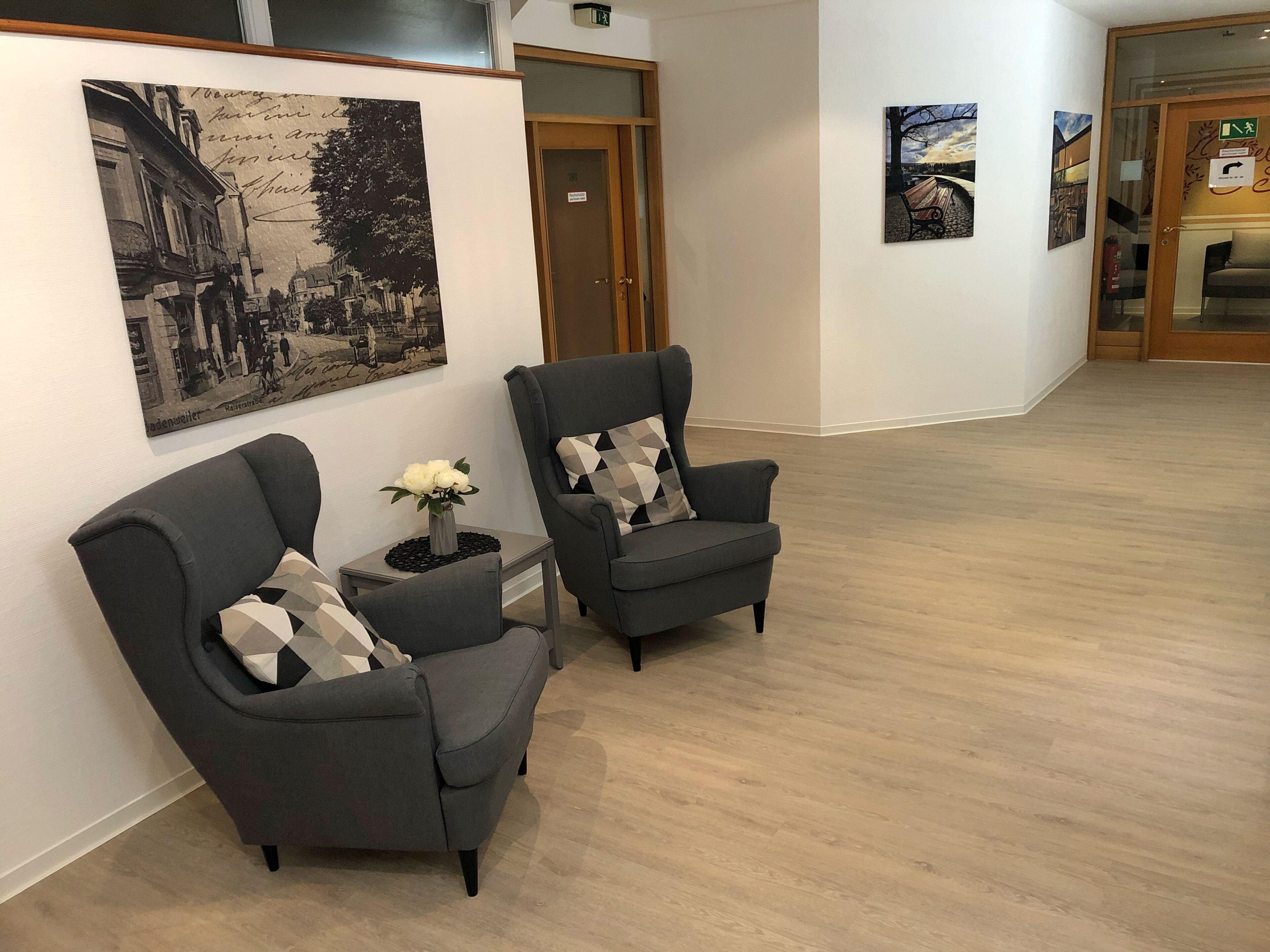 Two grey armchairs with patterned cushions, a small table with a vase of white flowers, and a black-and-white cityscape painting on the wall.