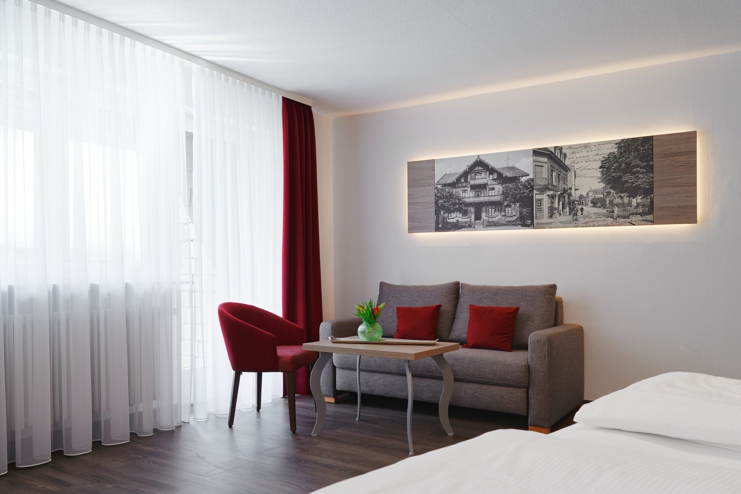 Habitación de hotel moderna con un sofá gris, una silla roja y una pequeña mesa con una planta. Un cuadro grande enmarco la pared.