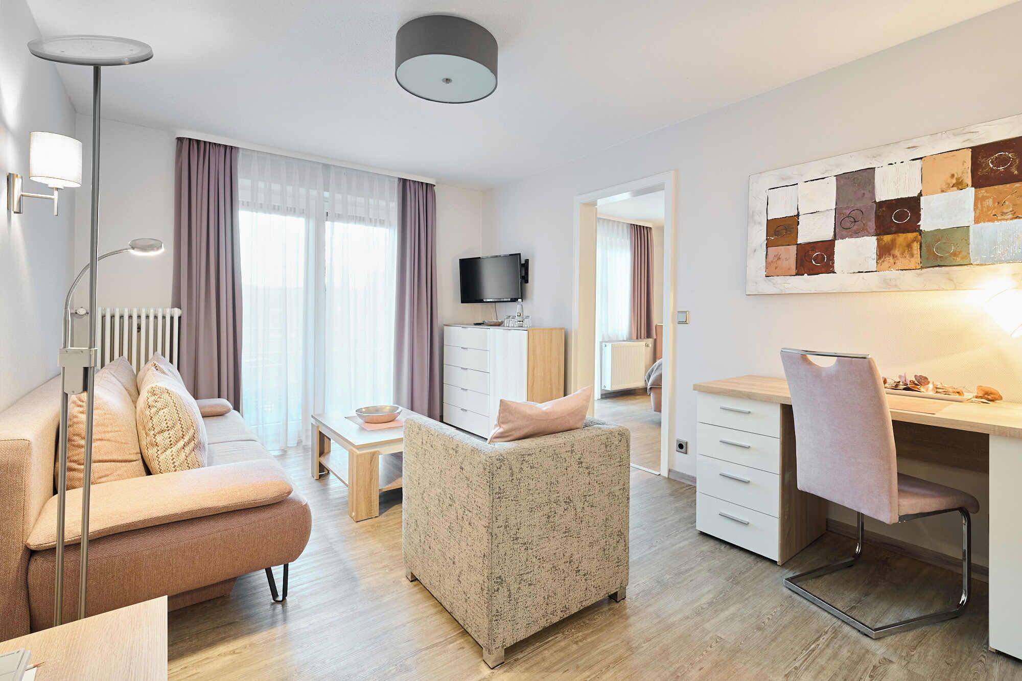 A modern hotel room with a sofa, a small table, a desk with a chair, a TV, and a painting on the wall.