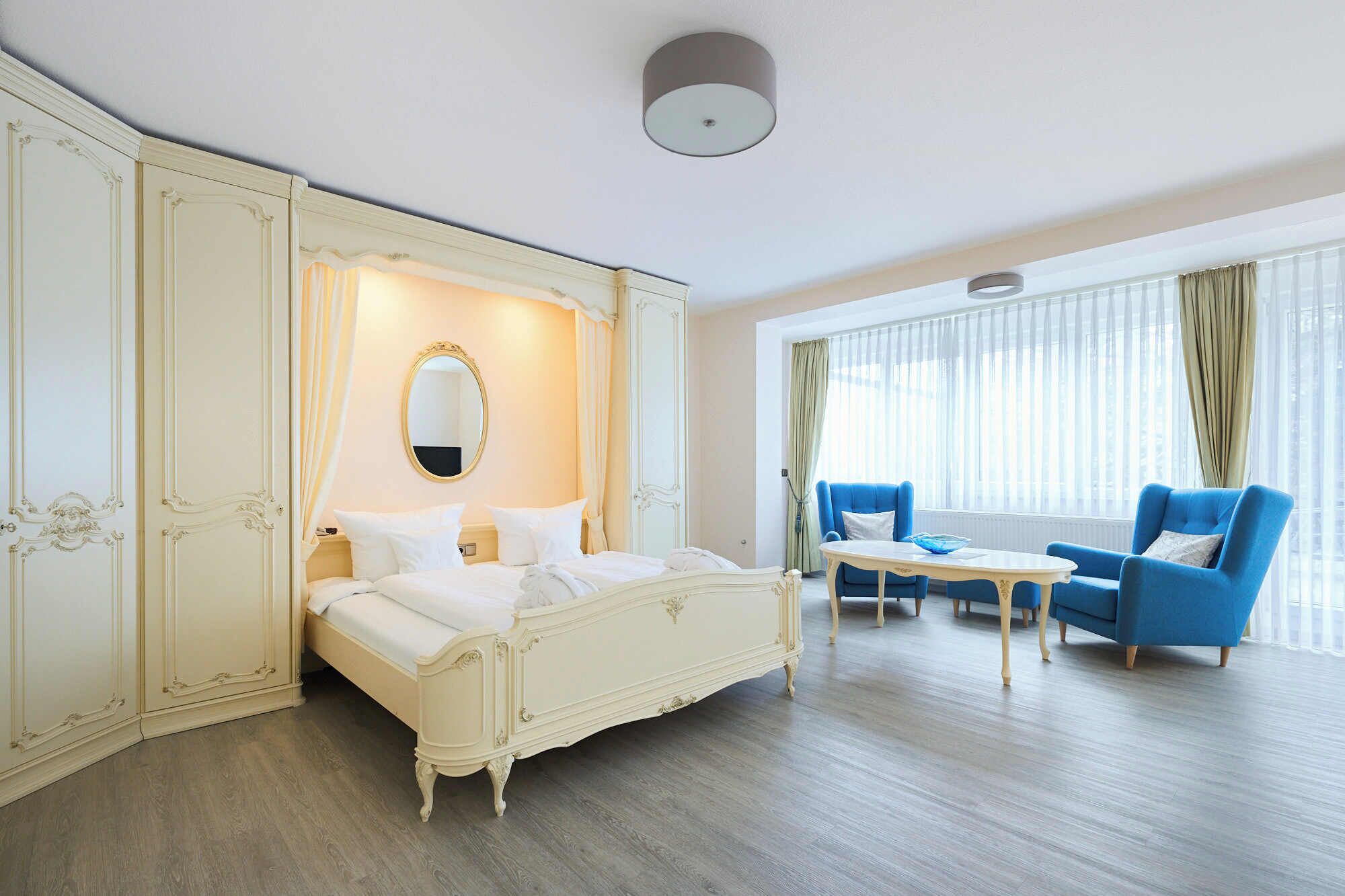A hotel room with a large bed, a sitting area with two blue armchairs, a small table, and a large window with curtains.
