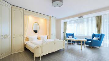 A hotel room with a large bed, a sitting area with two blue armchairs, a small table, and a large window with curtains.