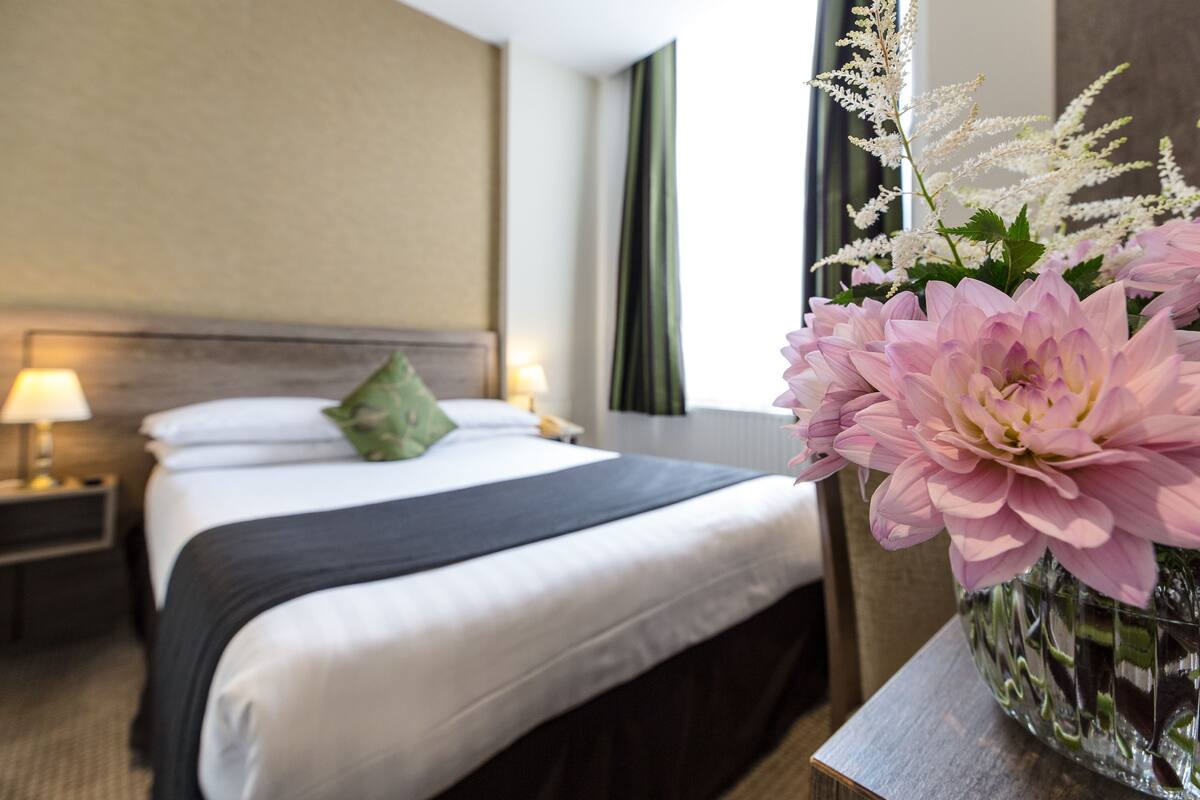Una habitación de hotel con cama, mesita de noche, lámpara, un florero con flores y una ventana con cortinas.