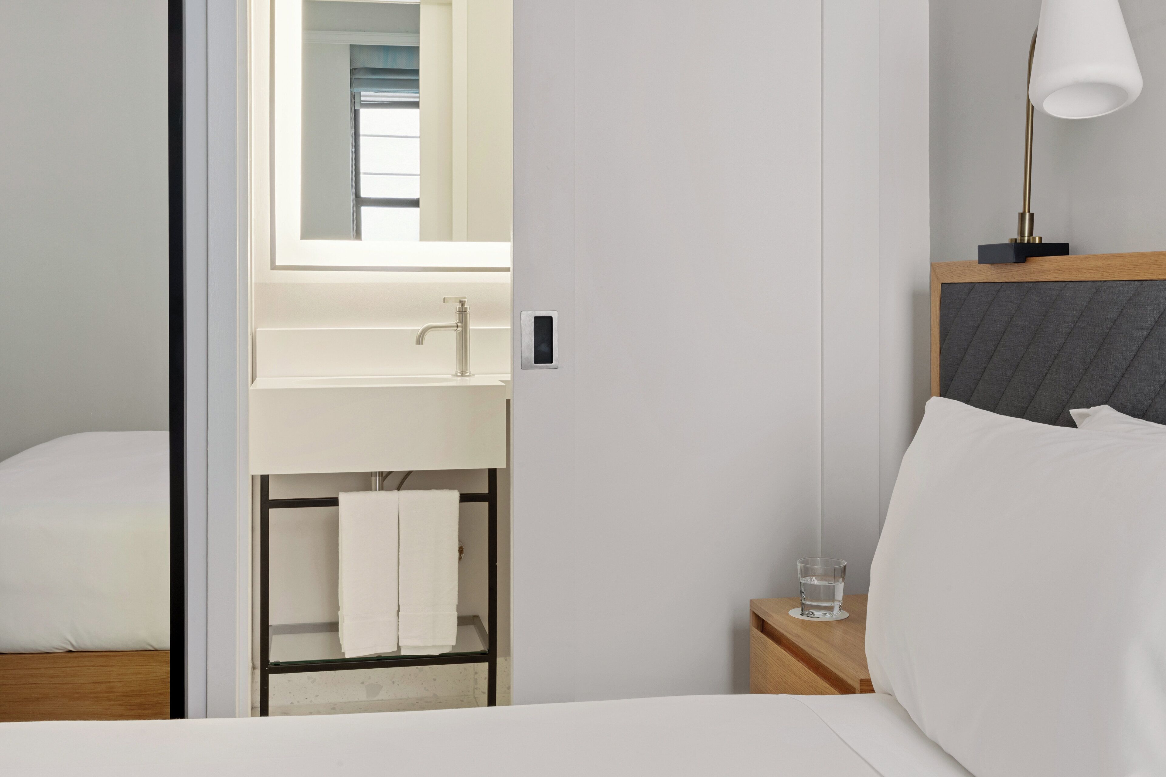 Ein Hotelzimmer mit Bett, Nachttisch, einem Tisch mit Lampe, einem Spiegel und einem Badezimmer mit Waschbecken und Handtuchhalter.