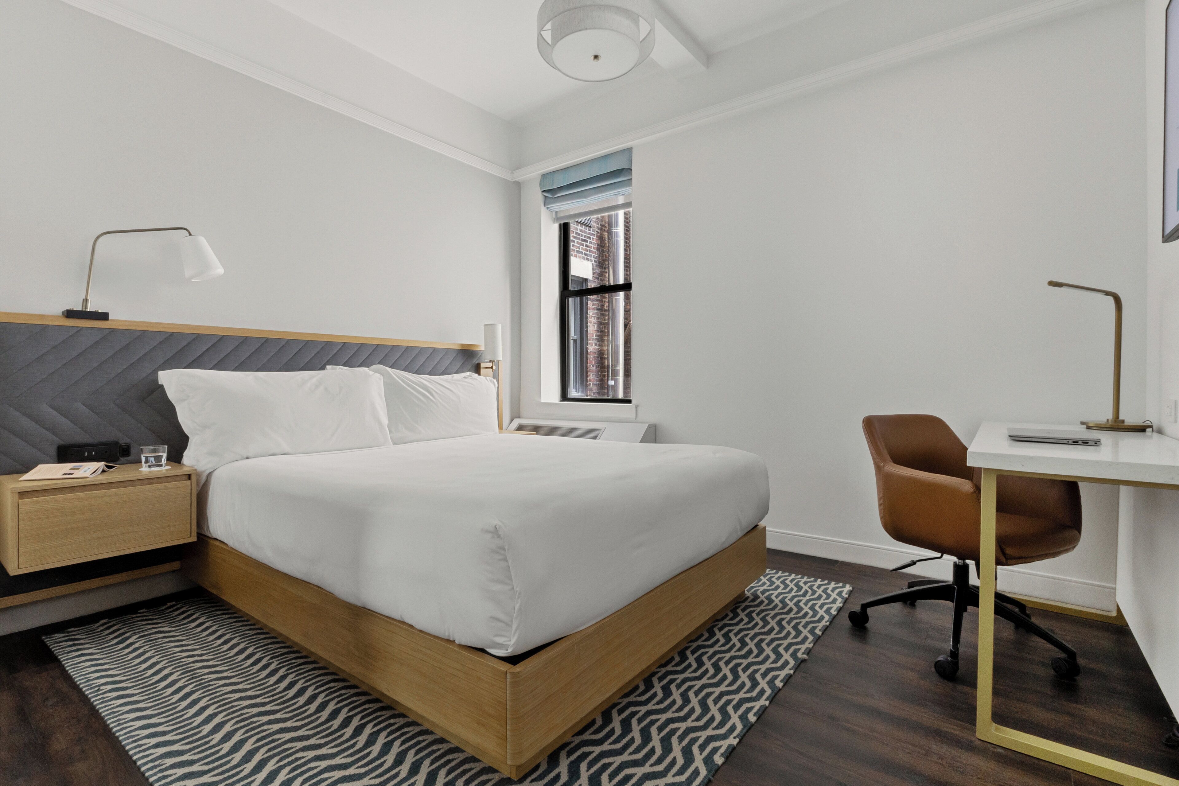 A modern hotel room with a bed, bedside table, lamp, desk, chair, and a window with a view of a brick building.