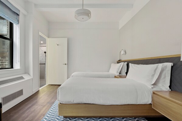 A modern bedroom with a large bed, bedside tables, a window with blinds, and a ceiling light.