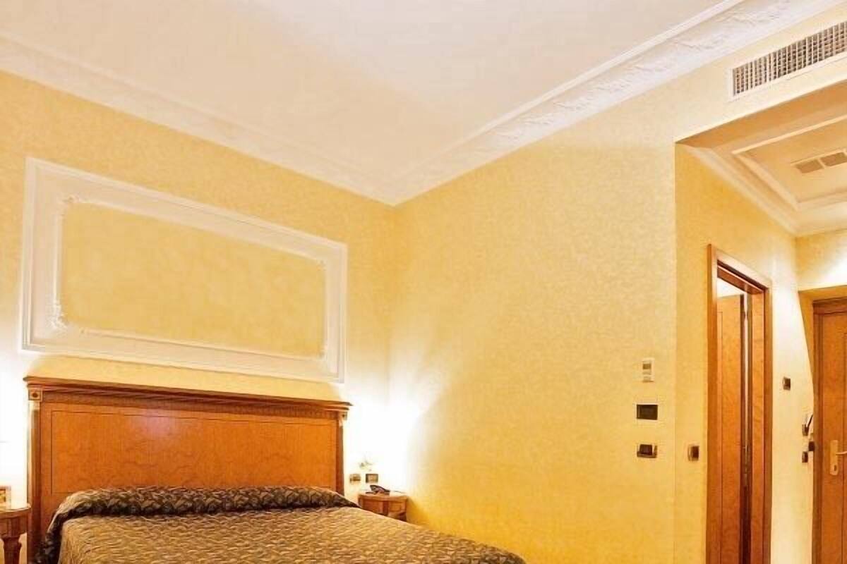 Habitación de hotel con una cama grande, un cabecero de madera, suelo alfombrado y una puerta a la derecha.