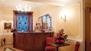 Un hall d’hôtel comprenant un comptoir d’accueil, un lustre, un miroir décoratif et une table avec des chaises.