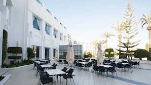 An outdoor dining area with tables and chairs, surrounded by a white building with large windows and a modern design. - Mahdia Palace Thalasso (Mahdia)