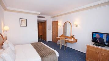 Quarto de hotel com uma cama, uma escrivaninha com espelho, uma televisão e uma porta que dá para outro quarto.
