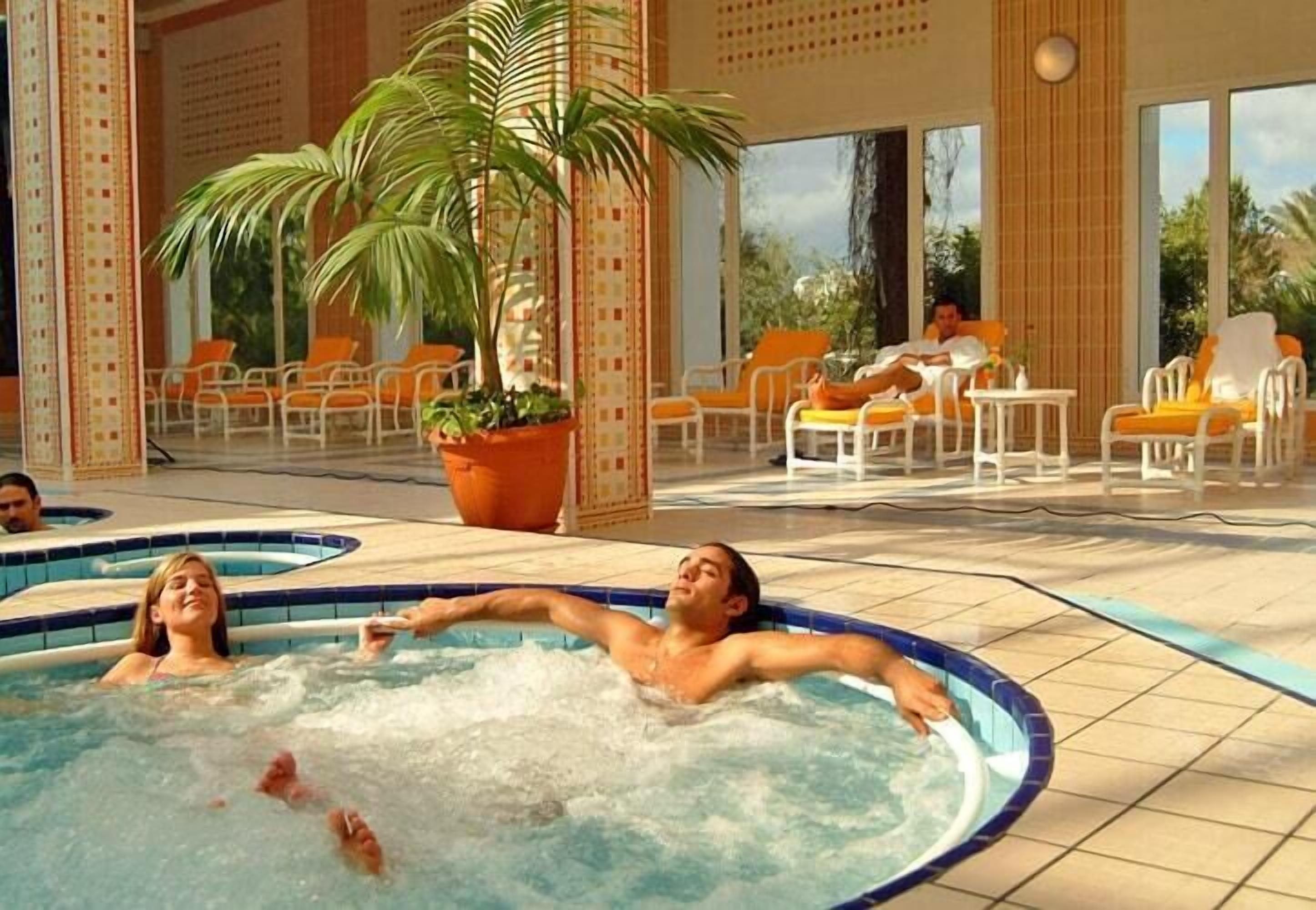 Zona de piscina con múltiples jacuzzis, rodeada de sillas anaranjadas y plantas en macetas.