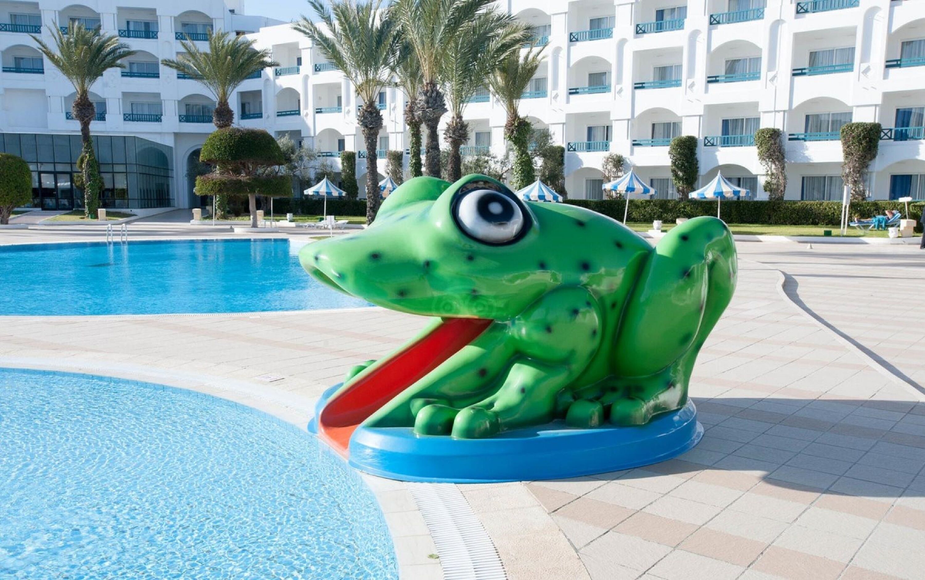A large green frog sculpture by a swimming pool with a hotel building in the background.