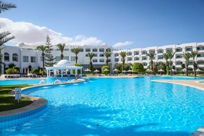Une grande piscine avec un kiosque, des palmiers et, au fond, un immeuble d’hôtel à plusieurs étages.