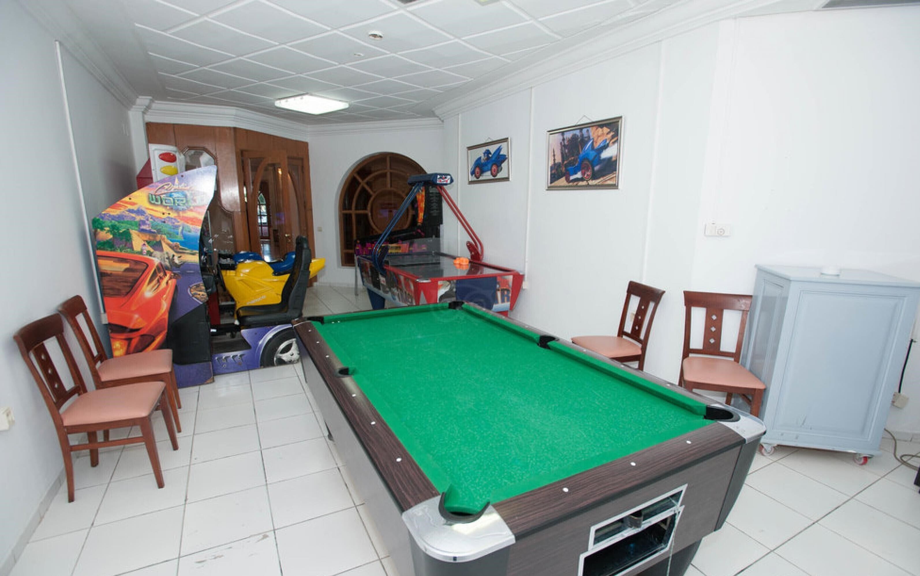 Una mesa de billar en una habitación con cuatro sillas de madera, una máquina recreativa colorida y dos cuadros en la pared.