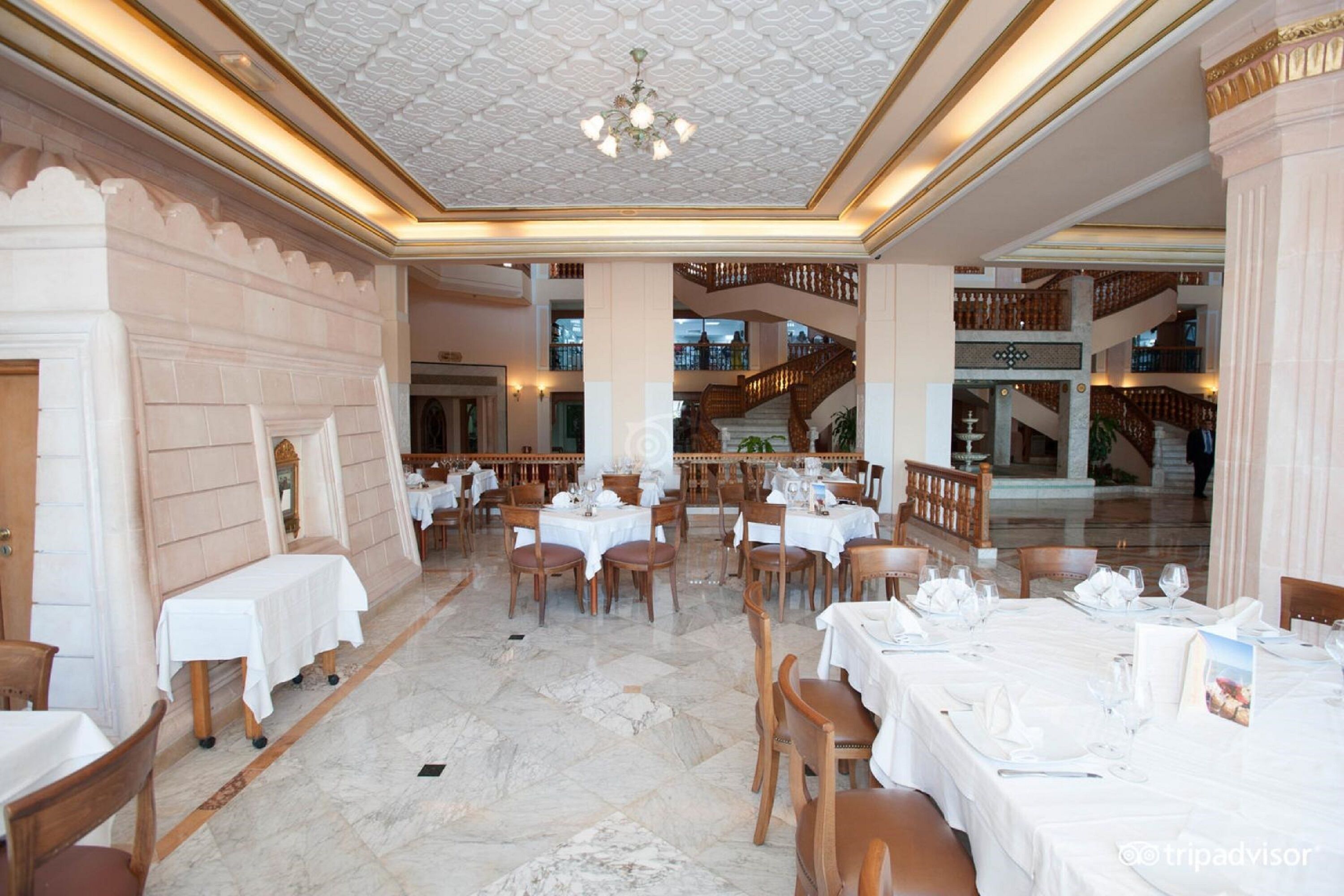 A spacious dining area with marble floors, multiple tables set for dining, and a grand staircase in the background.
