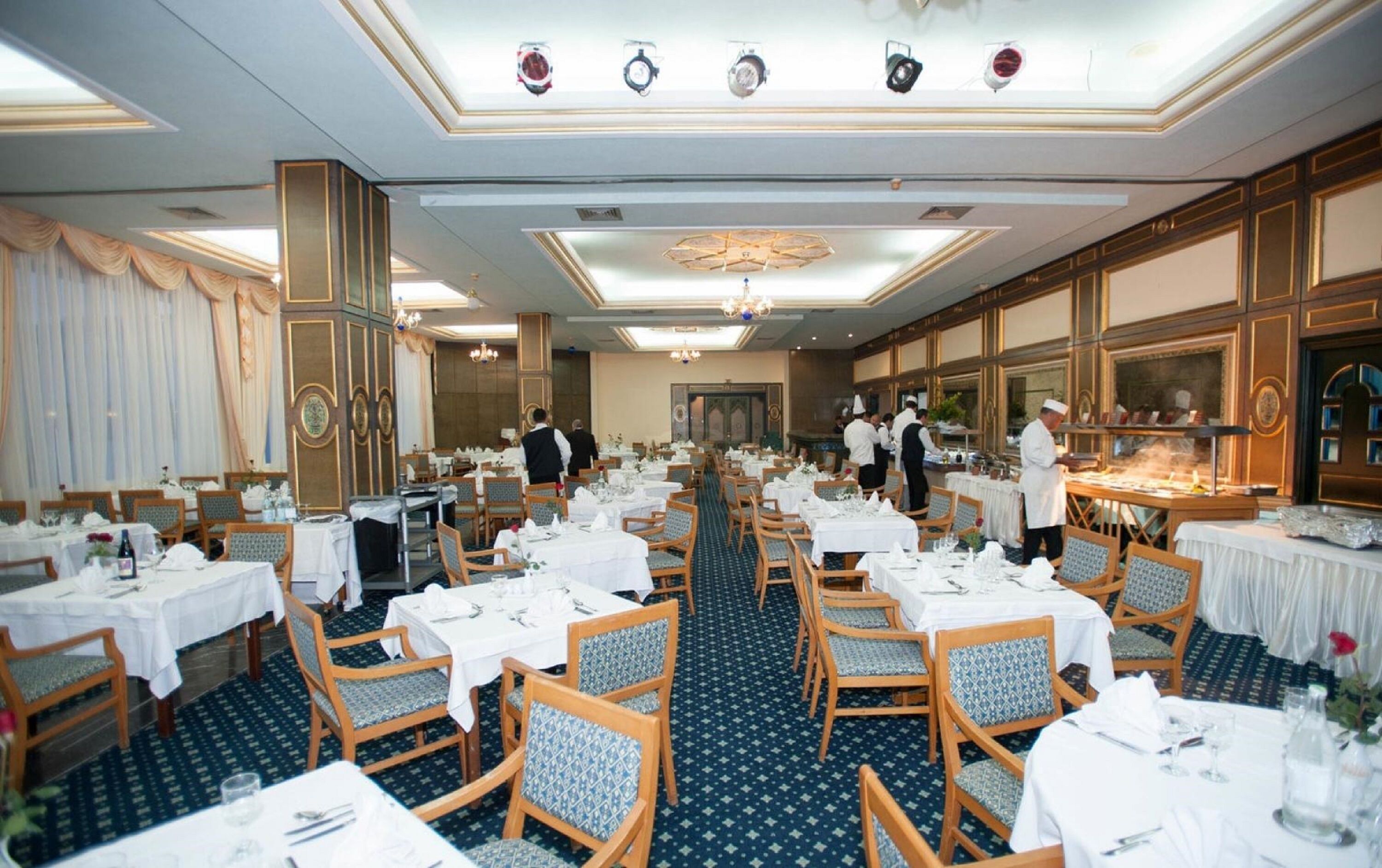 Un comedor formal con varias mesas, sillas de madera y una larga barra donde chefs preparan alimentos.
