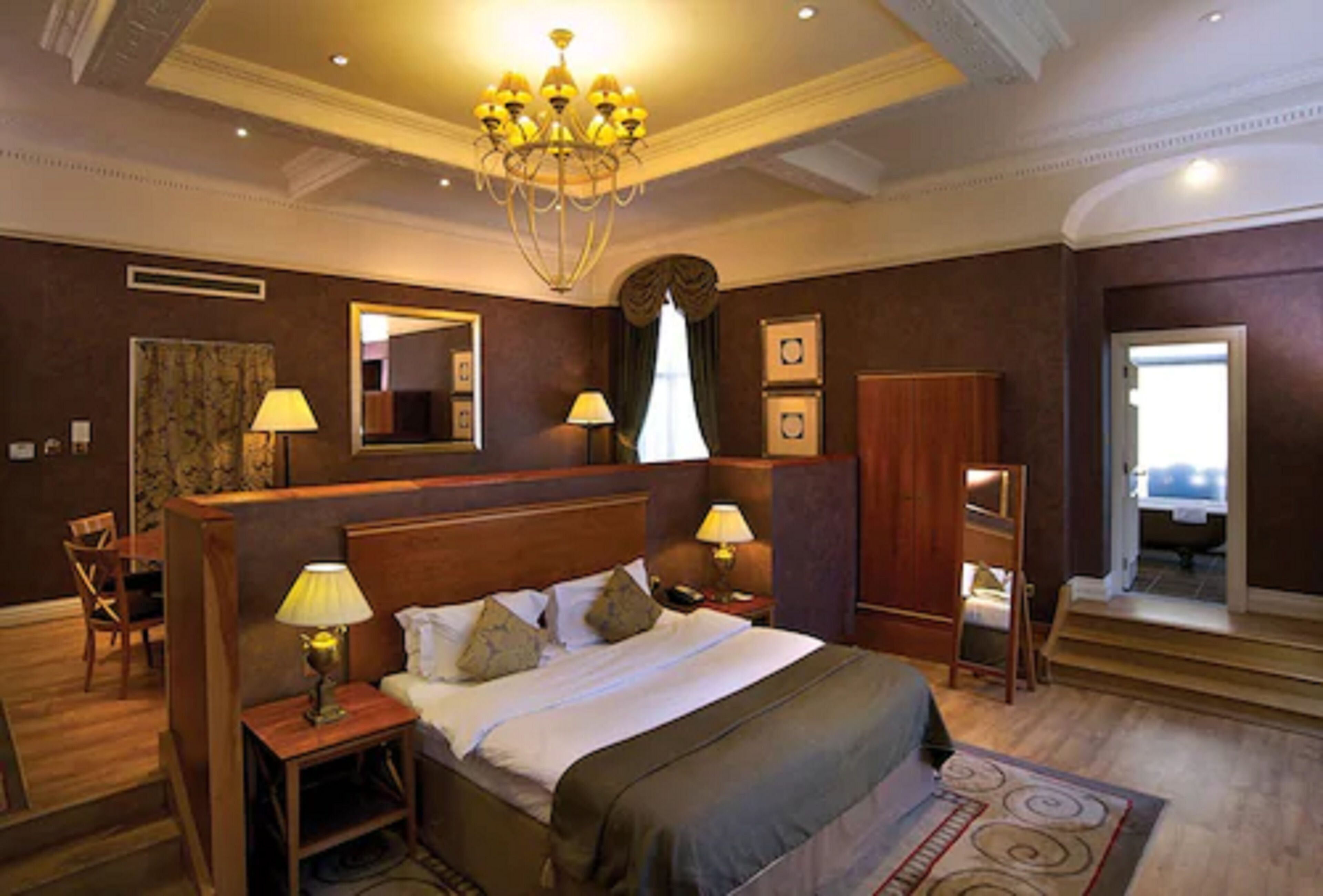 Ein Hotelzimmer mit einem großen Bett, zwei Nachttischen mit Lampen, einer Kristallleuchtre, einem Spiegel und Blick ins Freie.
