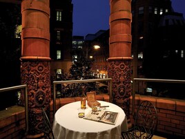 An outdoor dining area with a table set for two, surrounded by ornate columns, a Christmas tree, and a cityscape in the background.