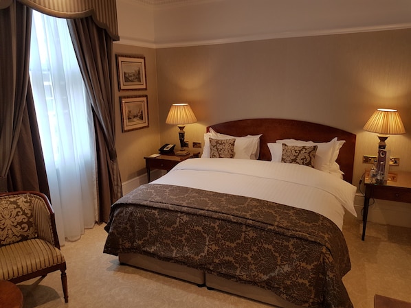 A hotel room with a large bed, two bedside lamps, a nightstand, a chair, and a window with curtains.