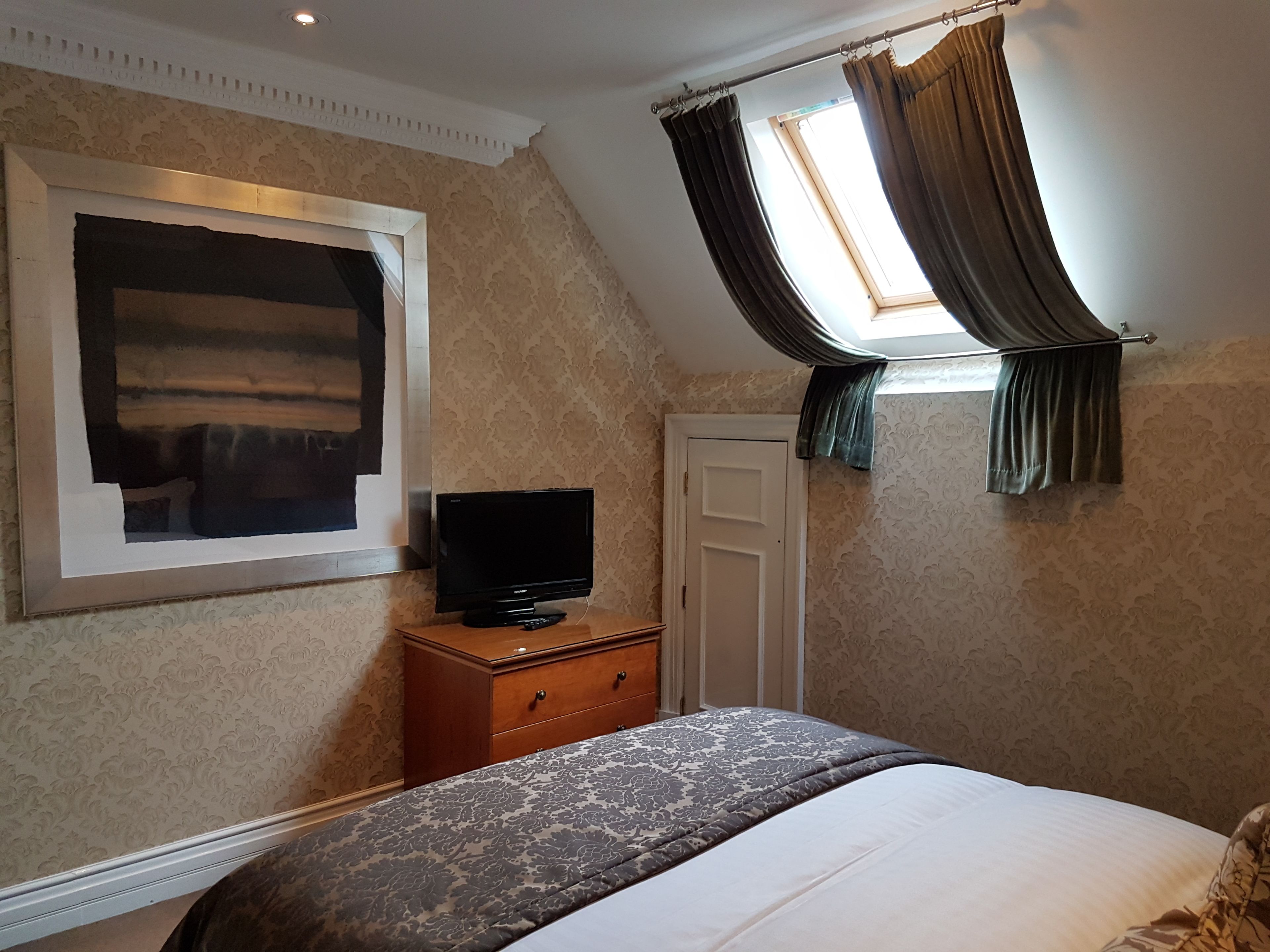 Un dormitorio con cama, televisor sobre una cómoda de madera, ventana con cortinas y pared con diseño.