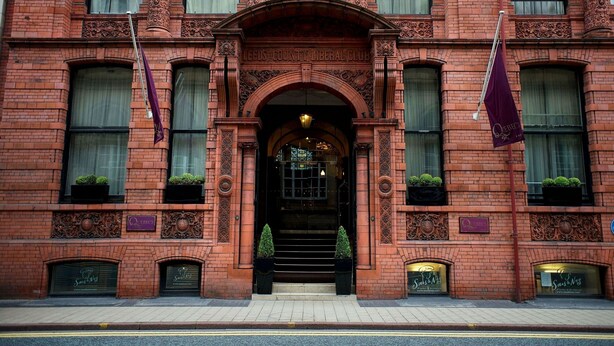 A red brick building with ornate architectural details, a central entrance with a canopy, and two flags. - Quebecs (Leeds)