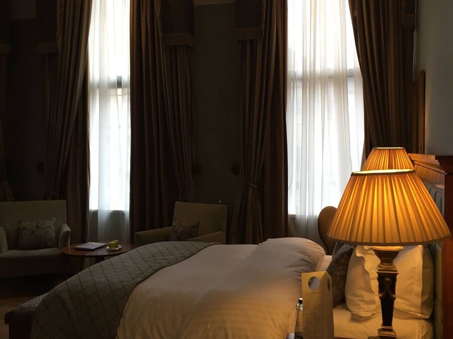 A hotel room with a bed, a bedside table with a lamp, a sofa, and two windows with curtains.