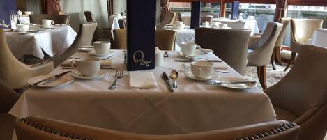 A dining area with tables set for four, each with white tablecloths, white plates, and silverware. There are brown upholstered chairs with a beaded trim. A dark blue menu stand is in the center of the tables.