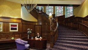 A grand lobby with a wooden reception desk, a purple armchair, and a staircase with stained glass windows. - Quebecs (Leeds)