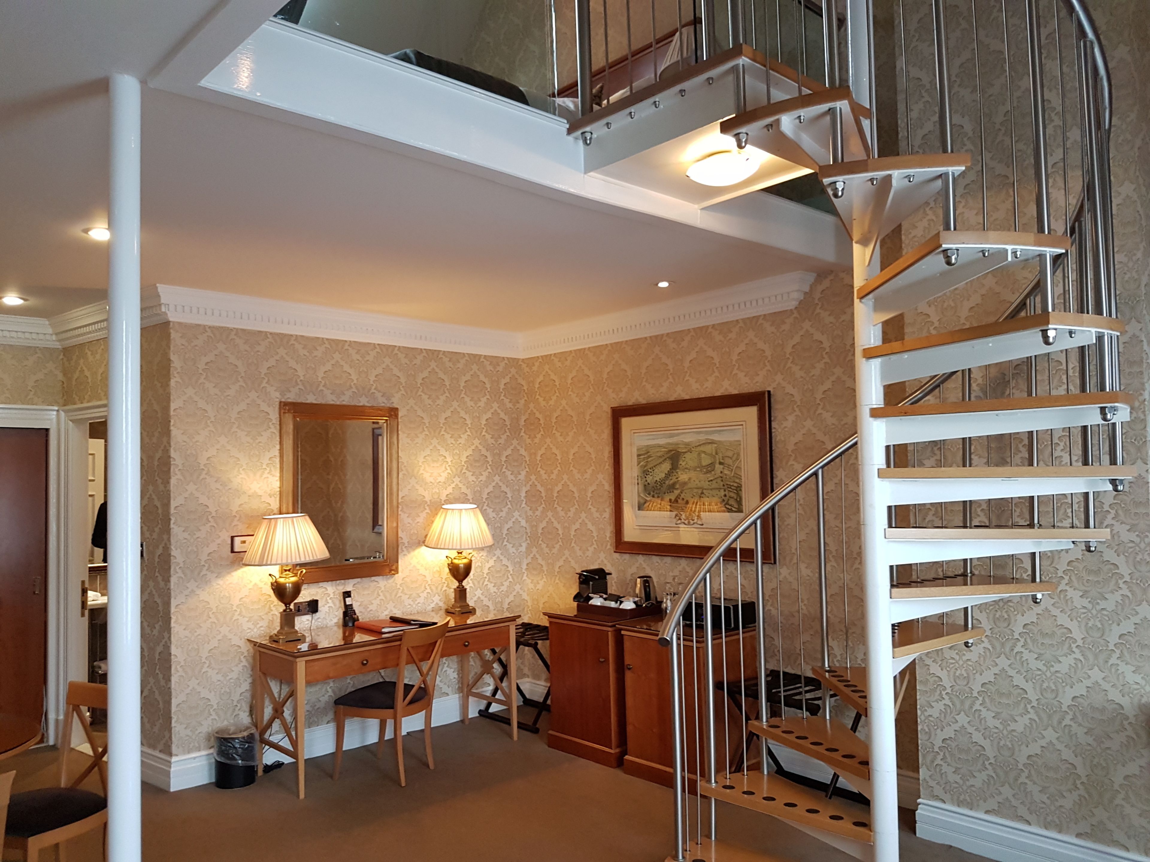 Une pièce avec un escalier en colimaçon, un bureau avec deux lampes, une chaise et un tableau encadré au mur.