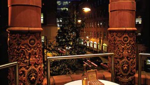 An outdoor dining area with ornate columns, a table set with a bottle, glasses, and a menu, and a view of a Christmas tree and buildings in the background. - Quebecs (Leeds)