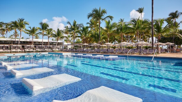 A swimming pool with lounge chairs and umbrellas, surrounded by palm trees. - Hotel Riu Palace Macao - Adults Only - All Inclusive (Punta Cana)