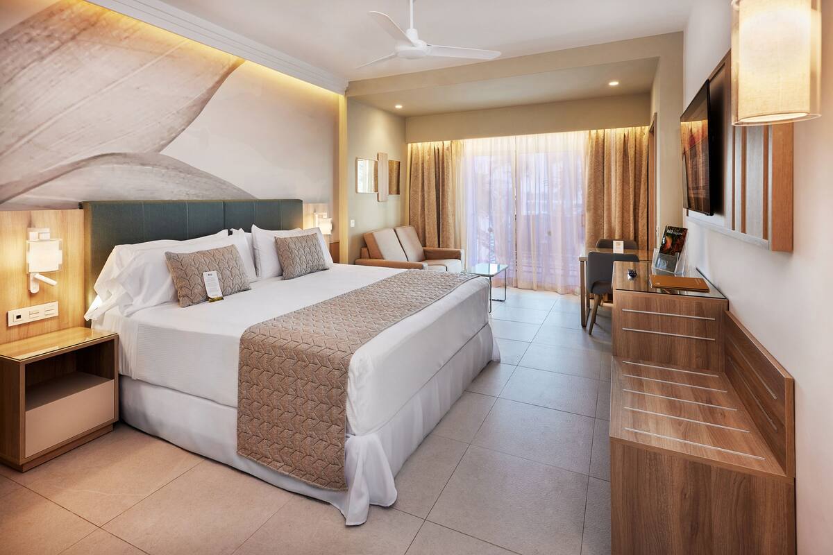A modern hotel room with a large bed, a desk, a chair, and a TV.