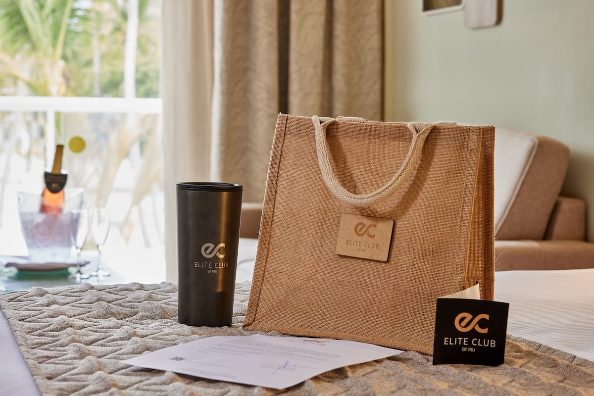 A hotel room with a bed, a bottle of Elite Club by RUI, a small black sign, and a textured area rug.